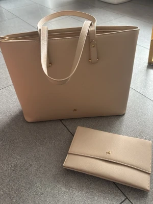 2 Beige väskor från Anna Field - Stilren beige handväska från Anna Field i slätt konstläder med matchande clutch. Väskan har två axelband, guldfärgade detaljer och en enkel logga framtill. Perfekt för dig som gillar minimalistisk design och vill ha plats för allt du behöver.