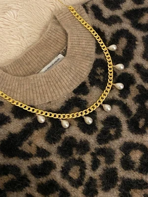 Guldfärgat halsband med pärlor - Snyggt guldfärgat halsband med chunky kedja och droppformade vita pärlor som hänger längs kedjan. Perfekt för att ge din outfit en cool och trendig touch. Materialet är rostfritt och pärlor, och designen är modern med en lyxig känsla.