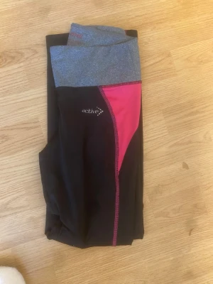 Svarta leggings med grå och rosa detaljer - Snygga svarta leggings från Active med bred grå midjeresår och en rosa panel längs sidan. Perfekta för träning eller chill, med bekväm passform och stretchigt material. Logga på låret och kontrastsömmar i rosa för extra stil.