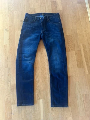Mörkblå skinny jeans från Lee - Säljer ett par mörkblå Daren Zip Fly  jeans från Lee med snygga slitningar på framsidan. Jeansen har en rak passform och Lee-logga på bakfickan. Perfekta för dig som gillar en stilren och modern look. Storlek W32 L32.