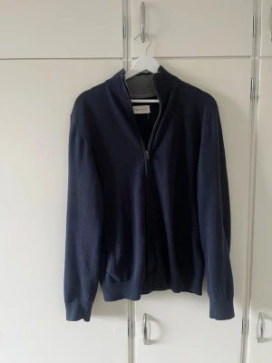 Mörkblå ziptröja  - Stilren mörkblå ziptröja med hög krage och hel dragkedja framtill. Tröjan har ribbade muddar vid ärmslut och nederkant, perfekt för en clean och enkel look. Klädesplaggets storlek är L men den sitter som en M.