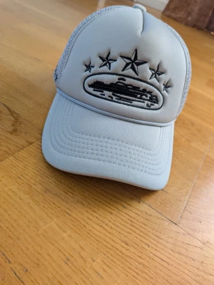 Ljusgrå truckerkeps med stjärnor - Snygg ljusgrå truckerkeps med svart broderi av stjärnor och ett motiv framtill. Kepsen har mesh på sidorna för extra ventilation och broderade stjärnor även på sidan. Perfekt accessoar för en cool streetstyle.