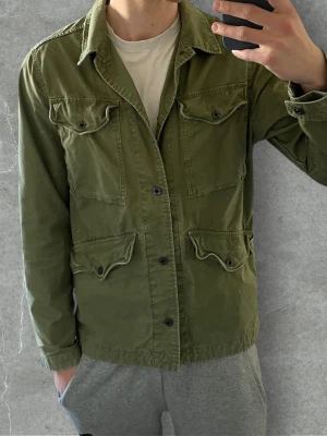 Grön utilityjacka från G-Star RAW | M - Snygg militärgrön utilityjacka från G-Star RAW med fyra fickor framtill och knäppning med svarta knappar. Jackan har klassisk krage och raka ärmar, perfekt för lager-på-lager-stil. Tillverkad i bomull för en bekväm känsla.