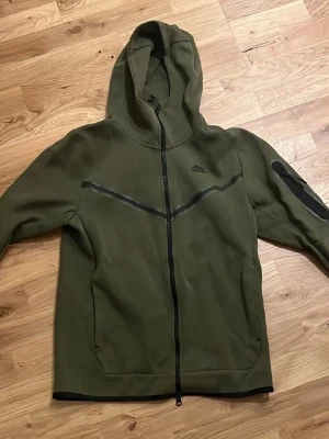 Olivgrön Nike hoodie med dragkedja - Snygg olivgrön hoodie från Nike med hel dragkedja och huva. Har svarta detaljer längs bröstet och på ärmen, samt fickor framtill. Perfekt för en avslappnad och sportig stil.
