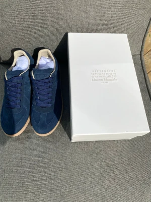 Maison Margiela blå sneakers i mocka - Snygga blå sneakers från Maison Margiela i mocka med beige detaljer och gummisula. Klassisk low-top modell med blå skosnören och diskret branding. Perfekta för dig som gillar stilrena och trendiga skor med premiumkänsla.