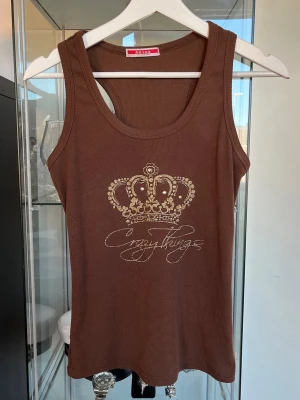 Brun ribbad linnetopp från Adina - Snygg brun ribbad linnetopp från Adina med ett glittrigt kronmotiv och texten 'Crazythings' framtill i storlek S/M. Toppen är ärmlös och har en rund halsringning, perfekt för en trendig och avslappnad look. Saknas en detalj i kronan framtill därav det låga priset. Axelbredd: 26cm. Längd: 56cm. Byst: 37cm.