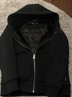 Moose knuckles bunny  - Snygg svart hoodie från Moose Knuckles med dragkedja framtill och stor huva. Insidan har ett quiltat, mönstrat foder och mjuk pälsimitation längs huvan. Stora muddar vid ärmslut och nederkant ger en schysst look. Perfekt för dig som gillar streetwear.