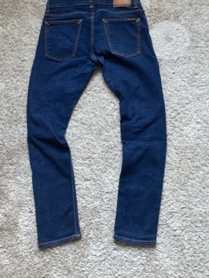 Mörkblå jeans från Nudie Jeans - Säljer ett par mörkblå jeans från Nudie Jeans med klassisk femficksdesign och kontrastsömmar. Jeansen har raka ben och en snygg läderpatch med logga bak i midjan. Materialet är slitstark denim i bomull. Perfekta för en clean och stilren look.