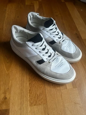 Sneaky Steve vita och grå sneakers  - Stilrena gats liknande sneakers från Sneaky Steve strlk 45 i vitt och ljusgrått med svarta detaljer. Skorna har klassisk låg modell, snörning och mocka detaljer. Perfekta för dig som gillar en clean och modern look.