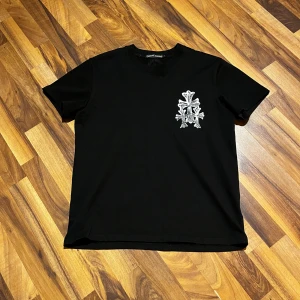 Svart t-shirt från Chrome Hearts - Snygg svart t-shirt från Chrome Hearts med ett coolt tryck i silver på bröstet och ryggen. Perfekt för dig som vill ha en stilren men ändå iögonfallande look. Passar bra till både jeans och shorts.