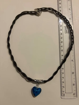 Halsband med blått hjärthänge - Snyggt halsband med tvinnad svart rem och ett blått hjärthänge i glas. Hänget har en cool mönstrad effekt och sitter fast i ett silverfärgat fäste. Perfekt accessoar för att lyfta din stil.