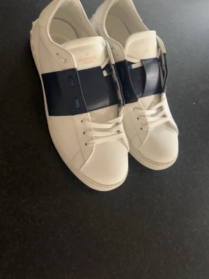Valentino opens - Snygga vita sneakers från Valentino med en bred mörkblå läderdetalj över sidan. Klassisk låg modell med vita snören och diskret logotyp på plösen. Yttermaterialet är läder och sulan har ett rutmönster för extra grepp.