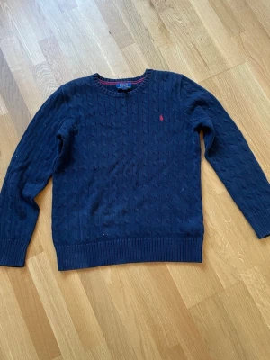 Mörkblå kabelstickad tröja Polo Ralph Lauren - Mörkblå kabelstickad tröja från Polo Ralph Lauren med rund halsringning och röd broderad logga på bröstet. Tröjan har ribbade muddar och är tillverkad i mjukt material. Perfekt för en stilren och klassisk look.