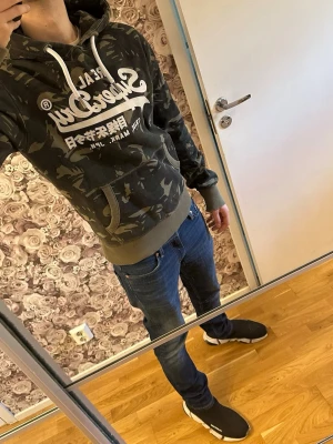  Superdry hoodie - Snygg hoodie från Superdry med camouflagemönster i gröna och svarta toner. Stor vit logga och japansk text på bröstet, samt vita snören i huvan. Perfekt för en chill och street-inspirerad stil.