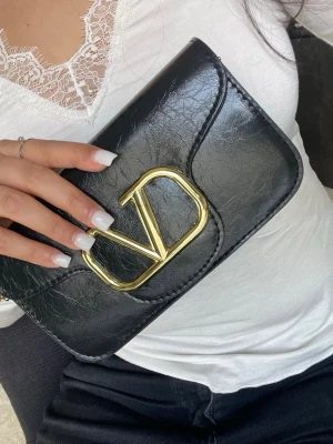 Svart axelväska i läderimitation - Snygg svart axelväska i läderimitation med enkel, stilren design och justerbar axelrem med guldfärgade detaljer. Perfekt storlek för det viktigaste och passar till många outfits. Lite missfärgning i spännet men det knappt syns.