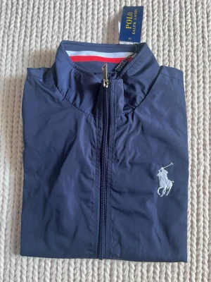 Marinblå vindjacka Polo Ralph Lauren - Snygg marinblå vindjacka från Polo Ralph Lauren med dragkedja framtill, broderad logga på bröstet och stor logotyp på kragen. Jackan har sportig känsla och ribbad krage med röd och vit detalj. Perfekt för en stilren och avslappnad look.