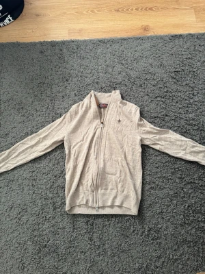 Beige ziptröja från Morris - Snygg beige ziptröja från Morris i 100% merinoull. Tröjan har en diskret logga på bröstet, ribbade muddar och hög krage. Perfekt för lager-på-lager och håller dig varm under kyliga dagar. Lite mindre i storlek 