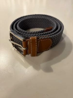 Grått flätat bälte med läderdetaljer - Snyggt grått flätat bälte med bruna läderdetaljer och silverspänne från revenite. Perfekt accessoar för att lyfta din outfit med en stilren och modern touch. Passar till både jeans och chinos.