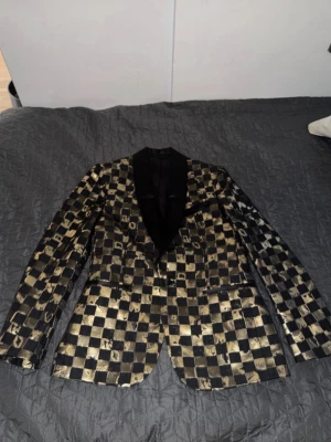 Svart och guld J.Lindeberg kavaj med schackmönster - Unik kavaj i svart och guld med schackrutigt mönster och svarta slag. Slimmad passform och glansigt material som sticker ut. Perfekt för dig som vill ha en statement-piece till garderoben. Stängs med en knapp framtill och har fickor på sidorna.
