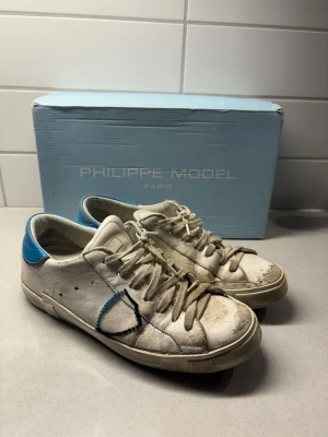 Philippe Model vita sneakers med blå detalj - Snygga vita sneakers från Philippe Model med blå detalj på hälen och broderad sköld på sidan. Klassisk låg modell med vita snören och diskret logga på plösen. Tillverkade i läder med slitstark gummisula. Perfekta för en clean och trendig look.