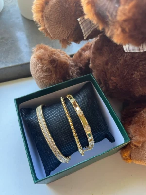  set med tre 18k guldpläterade, 16cm finns 17 och 18cm har bara ett exemplar passa på. - Snyggt set med tre armband i guldton. Ett armband har små glittrande stenar, ett är dekorerat med guldfärgade pärlor och det tredje har blommiga detaljer med stenar. Perfekt för att lyfta din stil med lite extra bling.