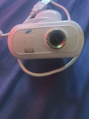 Vit USB-webbkamera 720p - Säljer en vit webbkamera med USB-anslutning och 720p-upplösning. Kameran har rund lins med silverfärgad kant och kompakt plastkropp. Perfekt för videosamtal och streaming.