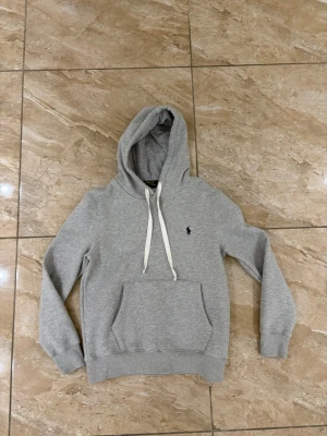 Grå hoodie från Polo Ralph Lauren - Snygg grå hoodie från Polo Ralph Lauren i slim fit-modell. Klassisk design med huva och vita dragsnören, känguruficka framtill och broderad logga på bröstet. Perfekt för en avslappnad och stilren look.