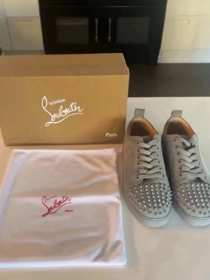 Grå Christian Louboutin sneakers med nitar 43 - Snygga grå sneakers från Christian Louboutin i mocka med silverfärgade nitar på tån och den ikoniska röda sulan. Skorna har vita skosnören och en låg siluett. Perfekta för dig som vill sticka ut med exklusiv streetstyle.