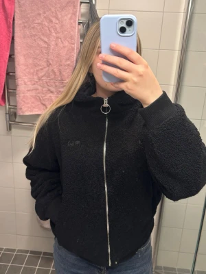 Svart teddyjacka från FILA med huva - Mysig svart teddyjacka från FILA i storlek M. Jackan har huva, dragkedja framtill med rund metallring och fickor med dragkedja. Perfekt för kyliga dagar och ger en chill vibe till din outfit.