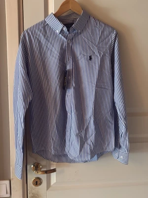 Blårandig skjorta från Ralph Lauren - Säljer en klassisk blårandig skjorta från Ralph Lauren med button-down krage och broderad logga på bröstet. Skjortan har lång ärm, knappar framtill och är i bomull. Perfekt för dig som gillar stilrena och tidlösa plagg. Endast använd en enda gång,riktigt bra skick 10/10. Hör av er vid eventuella frågor!