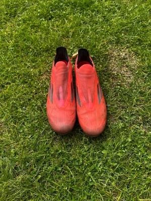 Röda Adidas X fotbollsskor - Säljer ett par röda Adidas X fotbollsskor med svarta detaljer och klassiska tre ränder på sidan. Skorna har låg vikt, strömlinjeformad design och är tillverkade i syntetmaterial för bästa grepp och känsla på planen. Perfekta för dig som vill ha snabbhet och kontroll.