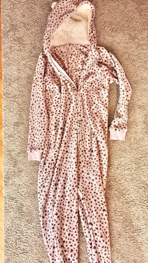 Rosa leopardmönstrad onesie med öron - Mysig rosa onesie med leopardmönster och huva med små öron. Materialet är mjukt fleece och insidan av huvan är vit och fluffig. Onesien har långa ärmar, dragkedja framtill och ribbade muddar vid ärmslut och benslut. Perfekt för att chilla hemma.