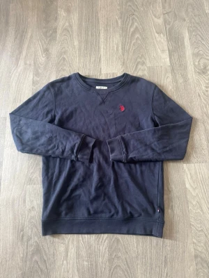 Mörkblå sweatshirt från polo assn - Mörkblå sweatshirt från polo assn med rund halsringning och diskret röd logobrodyr på bröstet. Tröjan har långa ärmar och ribbade muddar vid ärmslut och nederkant. Perfekt för en avslappnad och stilren look. Storleken är 12-13 men är större i storleken