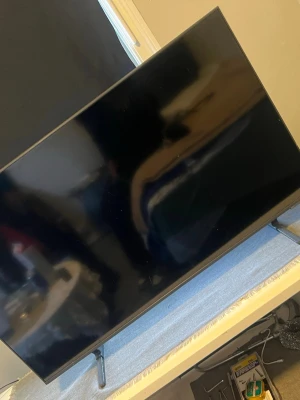 Svart platt TV med metallben - Säljer en stilren svart platt TV med tunna ramar och två metallben som stöd. Skärmen har en modern design och passar perfekt för gaming, streaming eller filmkvällar. TV:n har en rektangulär form och står stabilt på ett bord eller TV-bänk.