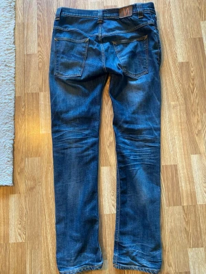 Mörkblå jeans från Nudie Jeans - Säljer ett par mörkblå jeans från Nudie Jeans med klassisk femficksdesign och raka ben. Jeansen har snygga slitningar och kontrastsömmar samt läderpatch med logga bak i midjan. Perfekta för en avslappnad och trendig look.