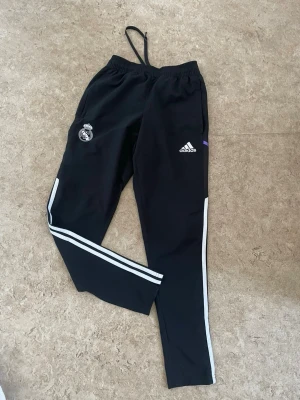 Svarta Adidas Real Madrid träningsbyxor - Säljer ett par svarta träningsbyxor från Adidas med Real Madrid-logga på ena benet och Adidas-logga på andra. Byxorna har vita ränder längs benen och snörning i midjan. Perfekta för träning eller fotbollspasset!