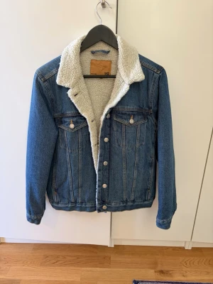 Blå jeansjacka med teddyfoder - Säljer en klassisk blå jeansjacka med vitt teddyfoder på insidan och kragen. Jackan har två bröstfickor med knappar och silverfärgade knappar framtill. Perfekt för dig som vill ha en tidlös och trendig look.
