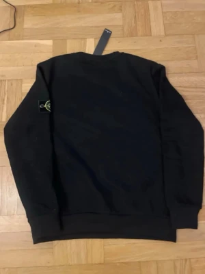 Svart sweatshirt från Stone Island - Svart sweatshirt från Stone Island med klassisk rund hals och ribbade muddar. Ikonisk logotyp-patch på vänster ärm. Perfekt för en stilren och avslappnad look.