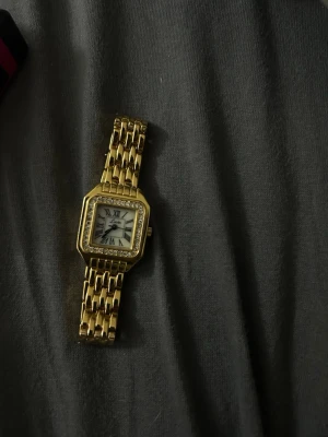 Guldfärgad fyrkantig klocka - Stilren guldfärgad klocka med fyrkantig urtavla och romerska siffror. Klockan har en länkarmband i metall och glittrande stenar runt urtavlan för extra bling. Perfekt accessoar för dig som vill sticka ut. (Kan få med justerare) 