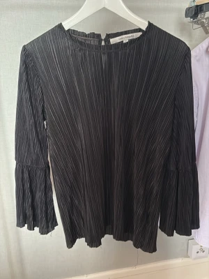 Svart plisserad blus från KappAhl - Trendig svart plisserad blus från KappAhl med rund halsringning och långa, utsvängda ärmar. Blusen har en snygg struktur och lätt glansigt tyg som ger en cool look. Perfekt att styla med jeans eller kjol för en modern vibe.