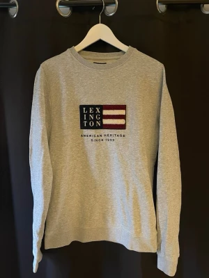Lexington tröja grå - Tja säljer en Snygg grå sweatshirt från Lexington med amerikanskt flaggtryck och text på bröstet. Tröjan har rund halsringning, långa ärmar och ribbade muddar. Perfekt för en avslappnad och stilren look. skick 10/10🤩har du några mer frågor eller funderingar är de bara att höra dig🤝