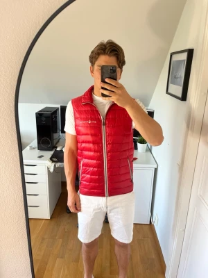 Röd dunväst från Sail Racing - Snygg röd dunväst från Sail Racing med hög krage och två dragkedjeförsedda fickor. Västen har quiltad design och logga på bröstet. Finns även två innerfickor, en på varsin sida. Hör gärna av dig vid frågor!
