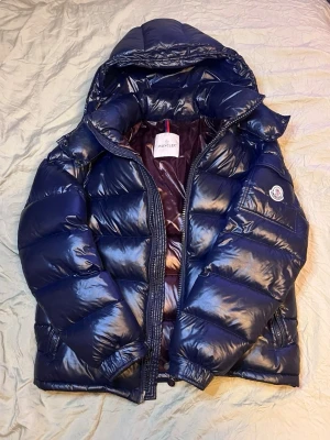 Mörkblå Moncler Maya  - Snygg mörkblå dunjacka från Moncler med glansig finish, stor huva och klassisk logga på ärmen. Jackan har breda kanaler, dragkedja framtill och två sidofickor. Perfekt för kalla vinterdagar och riktigt varm. Storlek 1/S inga defekter 