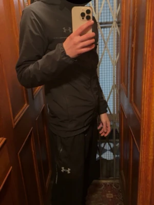 Svart träningsset från Under Armour - Säljer ett svart träningsset från Under Armour med matchande jacka och byxor. Setet har en sportig look med dragkedja på tröjan och diskret logga på byxan. Perfekt för gymmet eller löprundan. Byxorna - S, Jackan XS