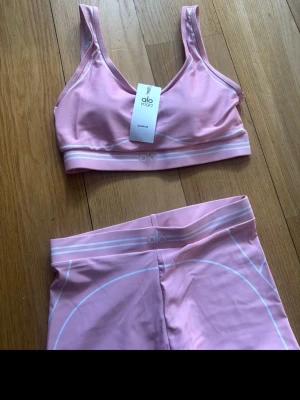 Rosa träningsset från Alo Yoga - Säljer ett rosa träningsset från Alo Yoga med sport-bh och matchande tights. Setet har vita detaljer och logga på både bh och byxor. Materialet är stretchigt och passar perfekt för yoga eller gym. Snygg och bekväm passform med bred resår i midjan.