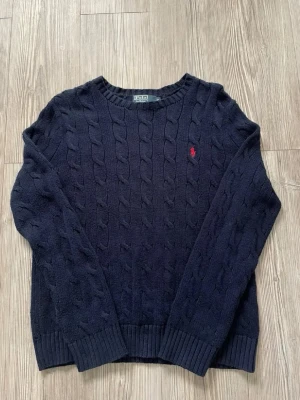 Mörkblå kabelstickad tröja Polo - Säljer en mörkblå kabelstickad tröja från Polo Ralph Lauren med klassisk rund hals och röd broderad logga på bröstet. Tröjan har ribbade muddar och är gjord i mjukt material som passar perfekt för lager på lager. Perfekt för dig som gillar stilrena plagg.