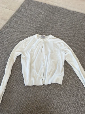Vit klassisk kofta med knappar - En stilren vit kofta med rund hals och knäppning framtill. Koftan har långa ärmar och ribbade muddar vid ärmslut och nederkant. Perfekt att slänga över en t-shirt eller topp för en enkel och clean look.