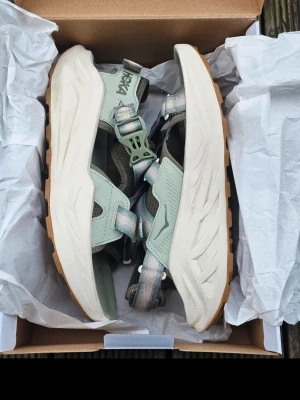 Hoka gröna sportiga sandaler - Säljer ett par sportiga sandaler från Hoka i ljusgrön färg med breda justerbara remmar och robust vit sula med brun yttersida. Sandalerna har en modern design och är tillverkade i syntetmaterial och mesh för extra komfort och ventilation.