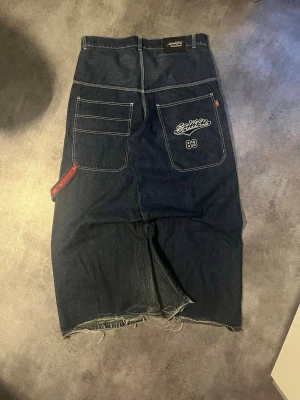 Baggy jeans JNCO style - Säljer ett par riktigt baggy jeans i mörkblå denim. De har stora bakfickor, broderad logga och nummer 83 på ena fickan samt råa, fransiga benslut. Perfekta för dig som gillar streetwear och loose fit.