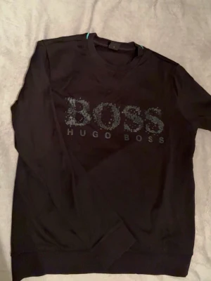 Svart sweatshirt från Hugo Boss - Svart sweatshirt med rund hals från Hugo Boss. Stor BOSS-logga framtill i en cool, texturerad design. Tröjan har långa ärmar och ribbade muddar vid ärmslut och nederkant. Perfekt för en avslappnad och stilren look.